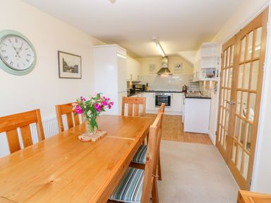 A dining area with a wooden table and chairs and a kitchen with white cabinets and appliances at Henrys Croft in Lizard