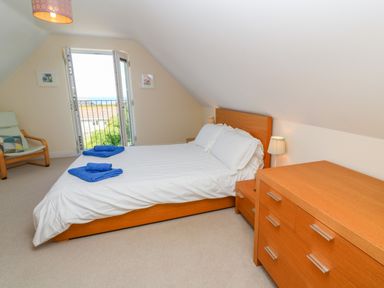 A bedroom with a double bed two blue towels a wooden dresser two bedside tables with lamps an armchair and a door opening to a balcony at Henrys Croft in Lizard