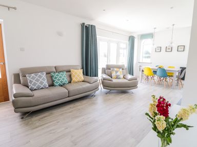 A living room with beige sofas and colorful cushions next to a dining table with multicolored chairs and flowers on a table at Senafe in Widemouth Bay
