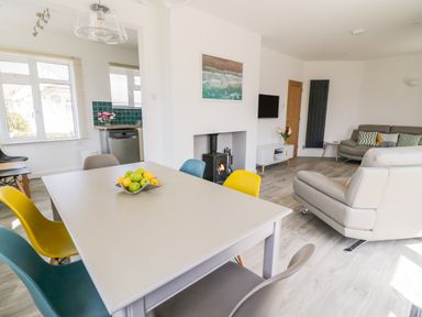 A dining table with colorful chairs and fruit bowl next to a living area with sofas and a fireplace at Senafe in Widemouth Bay