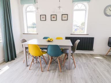 A dining room with a white table surrounded by yellow blue and grey chairs and two pendant lights at Senafe in Widemouth Bay