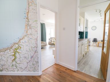 A hallway with wooden floor leading to a living room with armchairs and a kitchen with cabinets and a clock on the wall at Senafe in Widemouth Bay