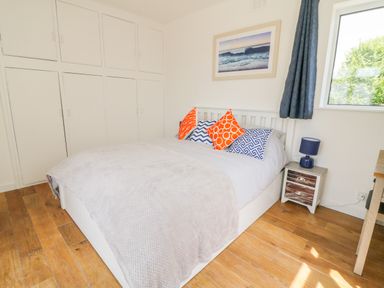 A bedroom with a bed with patterned pillows a nightstand with a lamp built in white cupboards and a window with curtains at Senafe in Widemouth Bay