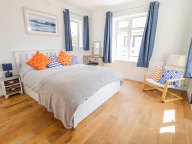 A bedroom with a white bed with colorful pillows a nightstand with a lamp a desk with a mirror a chair with pillows wooden floor and windows with blue curtains at Senafe in Widemouth Bay