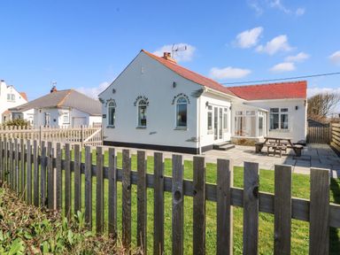 A white single-story house with a red roof and patio area behind a wooden fence at Senafe in Widemouth Bay
