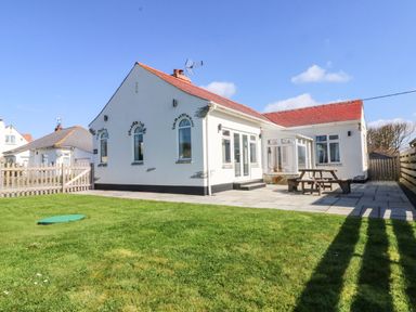 A white house with red roof tiles and arched windows next to a patio with a wooden picnic table and a grassy lawn at Senafe in Widemouth Bay