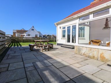 A paved patio with a wooden picnic table and benches next to a white house with large windows at Senafe in Widemouth Bay