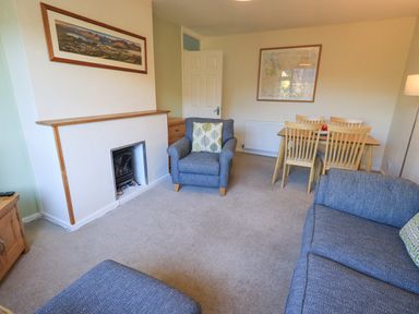 A living room with blue armchairs a dining table with wooden chairs a small fireplace and framed pictures on the walls at Fairfell in Keswick