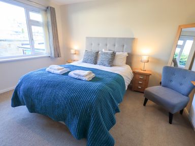 A bedroom with a bed with blue bedding two bedside tables with lamps a grey chair and a large window at Fairfell in Keswick
