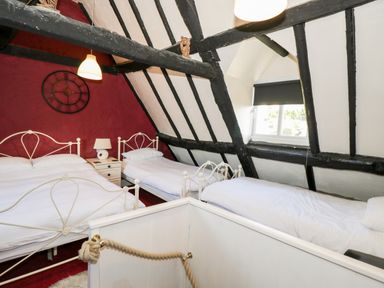 A bedroom with three white metal beds a nightstand with a lamp a wall clock window with blinds and exposed black beams at Combe Cottage in Brimscombe near Stroud