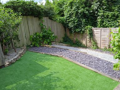 A garden with artificial grass lawn bordered by gravel and slate chips and wooden fences covered in green plants at Combe Cottage in Brimscombe near Stroud