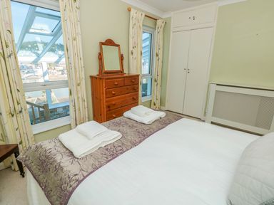 A bedroom with a bed, folded towels, wooden dresser with mirror, windows with curtains, and built-in cupboards at Estuary View House Annexe in Dartmouth