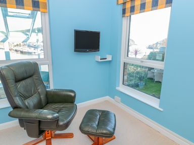 A corner room with a black leather chair and footstool a wall-mounted TV and windows showing water views at Estuary View House and Annexe in Dartmouth