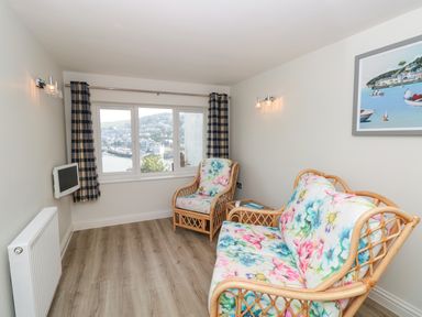 A small sitting room with floral cushions on rattan sofa and armchair a small wall-mounted tv and window with check curtains at Estuary View House and Annexe in Dartmouth
