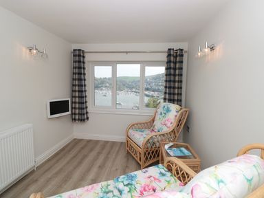 A small sitting room with floral cushioned wicker chairs a small glass table a wall-mounted television and a window with a view of a harbor at Estuary View (House & Annexe) in Dartmouth
