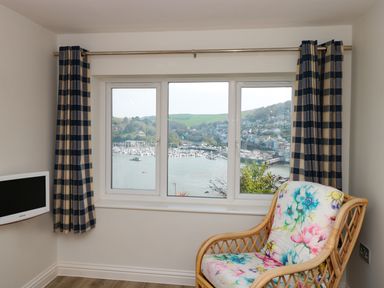 A window with a view of a harbor with boats and hills with striped curtains and a floral patterned chair at Estuary View House and Annexe in Dartmouth