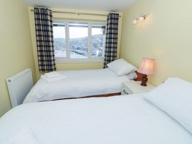 A bedroom with two single beds a nightstand with a lamp and a window with curtains overlooking a harbor at Estuary View House and Annexe in Dartmouth