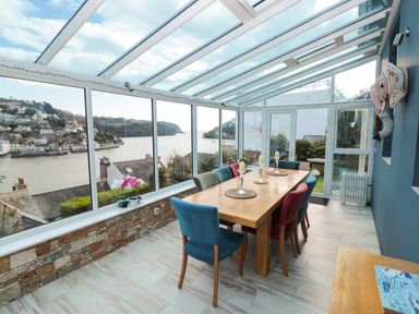 A dining area with a wooden table and colorful chairs in a glass conservatory overlooking a river and houses at Estuary View House and Annexe in Dartmouth