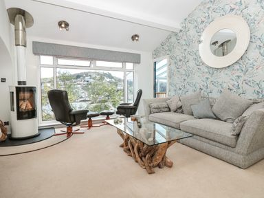 A living room with a gray sofa a glass coffee table with a wooden base two black chairs near large windows and a wall-mounted round mirror at Estuary View House and Annexe in Dartmouth