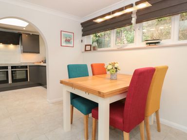 A dining area with a wooden table and four colorful chairs next to a kitchen with dark cabinets at Estuary View House and Annexe Dartmouth