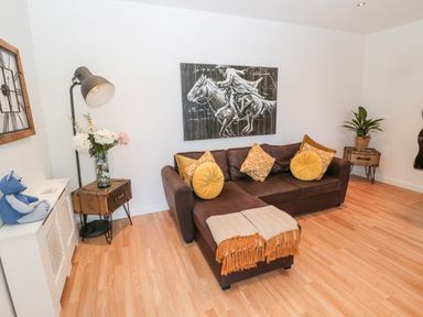 A living room with a brown sofa with yellow cushions and a blanket a floor lamp a side table with flowers a wall clock and a plant at The Snug in Beadnell