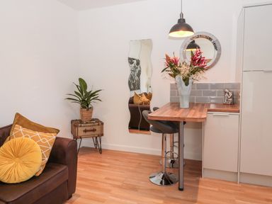 A small dining area with a wooden table, two black bar stools, a vase with flowers, a brown sofa with yellow cushions, a plant, and a wavy mirror on the wall at The Snug in Beadnell