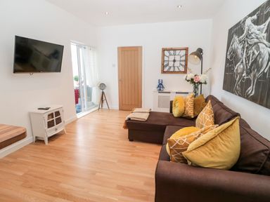 A living room with a brown sectional sofa with yellow cushions a wall clock a floor lamp a television on a stand and a sliding glass door at The Snug in Beadnell