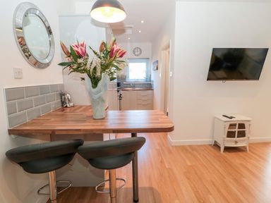 A kitchen area with a wooden breakfast bar and flowers in a vase with two bar stools a wall-mounted tv and a small cabinet on a wooden floor at The Snug in Beadnell