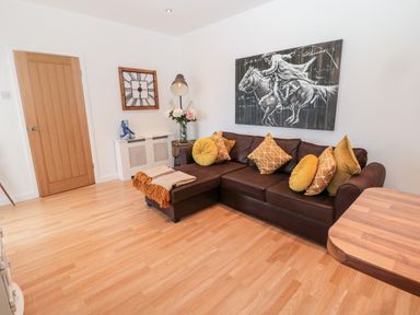 A living room with a brown sectional sofa with yellow and patterned cushions a wooden door a large wall clock a floor lamp and horse artwork at The Snug in Beadnell