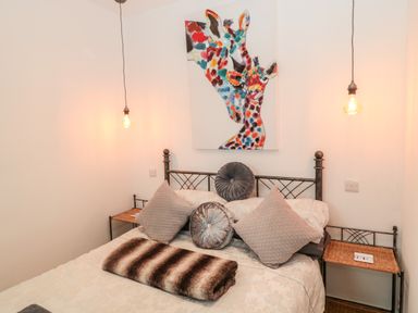 A bedroom with a metal bed frame two hanging light bulbs a colorful giraffe painting and patterned pillows at The Snug in Beadnell
