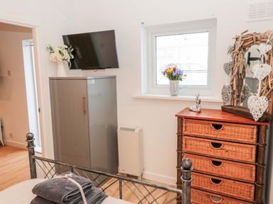 A bedroom with a metal bed frame folded towels on the bed a grey wardrobe with a TV above it a window with flowers on the sill and a wicker chest of drawers with a decorative mirror at The Snug in Beadnell