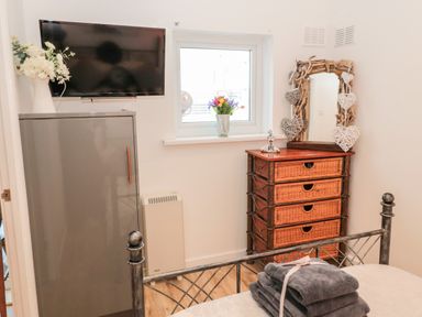 A bedroom with a wall-mounted television a gray cabinet a window a wicker drawer unit with a decorative mirror and folded towels on a metal bed frame at The Snug in Beadnell