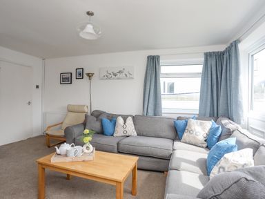 A living room with a gray sectional sofa, blue and patterned cushions, a wooden coffee table with a tray, a beige armchair, framed pictures, and blue curtains at Crows Nest - Rhosneigr in Rhosneigr