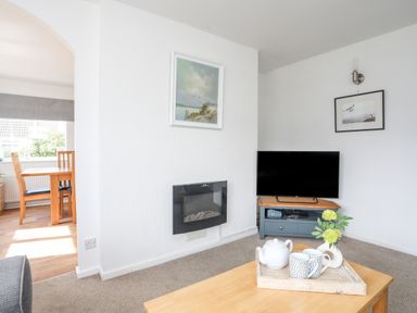 A living room with a wall-mounted electric fireplace a television on a stand wooden table with a tray teapot cups and a vase with flowers and a dining area with wooden chairs and table through an archway at Crows Nest - Rhosneigr in Rhosneigr