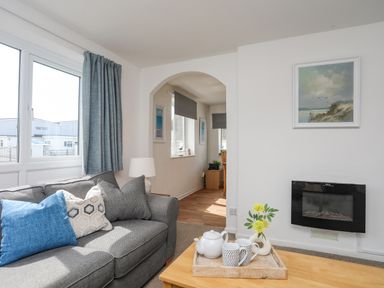 A living room with a grey sofa with pillows a wooden coffee table with a teapot cups and flowers a wall-mounted electric fireplace and a painting above it at Crows Nest - Rhosneigr in Rhosneigr