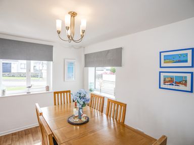 A dining room with wooden table and chairs near windows with grey blinds and wall art at Crows Nest - Rhosneigr in Rhosneigr