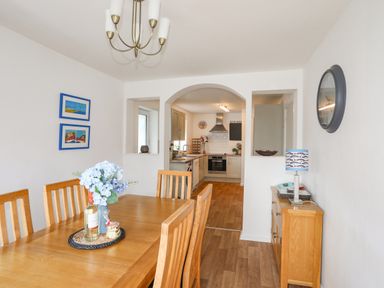 A dining room with a wooden table and chairs and a kitchen visible through an arched doorway at Crows Nest - Rhosneigr in Rhosneigr