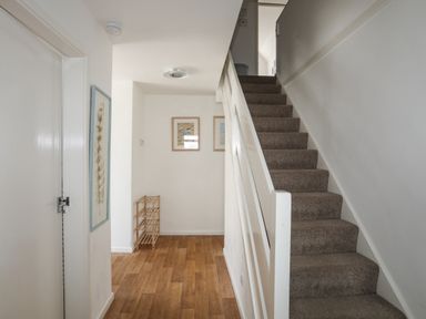 A hallway with carpeted stairs on the right wooden floor a closed door on the left and framed pictures on the wall at Crows Nest - Rhosneigr