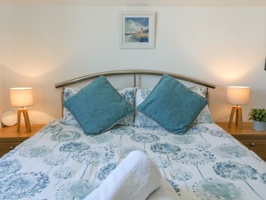 A bedroom with a bed with floral bedding two blue pillows two lamps on wooden bedside tables and a framed picture on the wall at Crows Nest - Rhosneigr