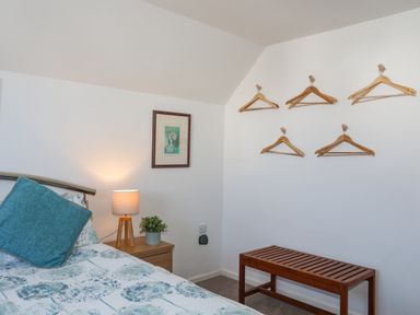 A bedroom with a bed featuring blue floral bedding and a teal pillow a wooden nightstand with a lamp and plant a framed picture on the wall wooden hangers on hooks and a wooden bench at Crows Nest - Rhosneigr in Rhosneigr