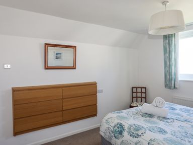 A bedroom with a bed with floral bedding a wooden dresser a wooden chair and a window with blue curtains at Crows Nest - Rhosneigr in Rhosneigr