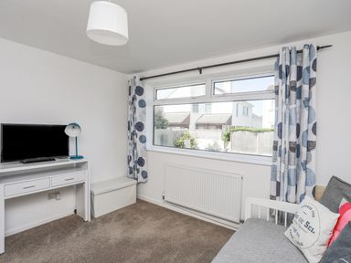 A room with a window with patterned curtains a white desk with a television and lamp a white storage bench and a gray sofa with pillows at Crows Nest - Rhosneigr in Rhosneigr