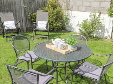 An outdoor garden with a round metal table and four chairs with cushions and two recliner chairs with pillows at Crows Nest - Rhosneigr in Rhosneigr