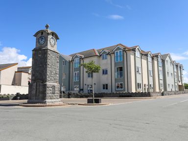 A clock tower near a multi-story residential building and a tree on a street at Crows Nest - Rhosneigr in Rhosneigr