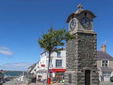A stone clock tower near a tree on a street with shops and houses by the sea at Crows Nest Rhosneigr