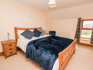 A bedroom with a wooden bed, blue bedding, pillows, wooden nightstand, window with plaid curtains at Gwynfa in Capel Iwan near Newcastle Emlyn