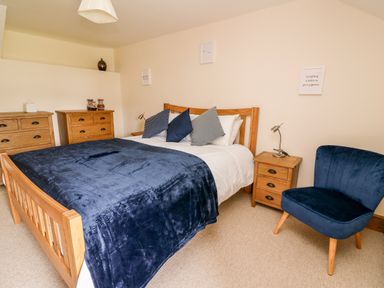 A bedroom with a wooden bed with blue bedding a wooden dresser and bedside table a blue chair and framed wall art at Gwynfa in Capel Iwan near Newcastle Emlyn