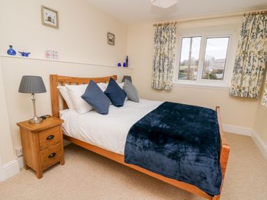 A bedroom with a wooden bed white bedding blue pillows a dark blue throw a wooden nightstand with a lamp and a window with patterned curtains at Gwynfa in Capel Iwan near Newcastle Emlyn