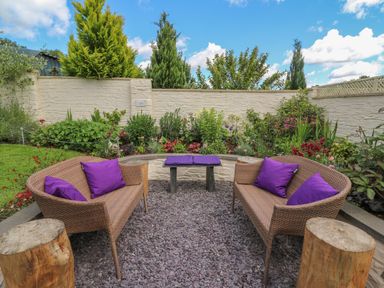 An outdoor seating area with two wicker sofas with purple cushions a small table and wooden stools surrounded by plants and a beige wall at Gwynfa in Capel Iwan near Newcastle Emlyn