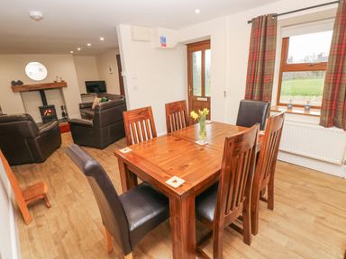 A dining area with wooden table and six chairs near a window and door connected to a living room with leather sofas and a wood stove at Gwynfa Capel Iwan near Newcastle Emlyn